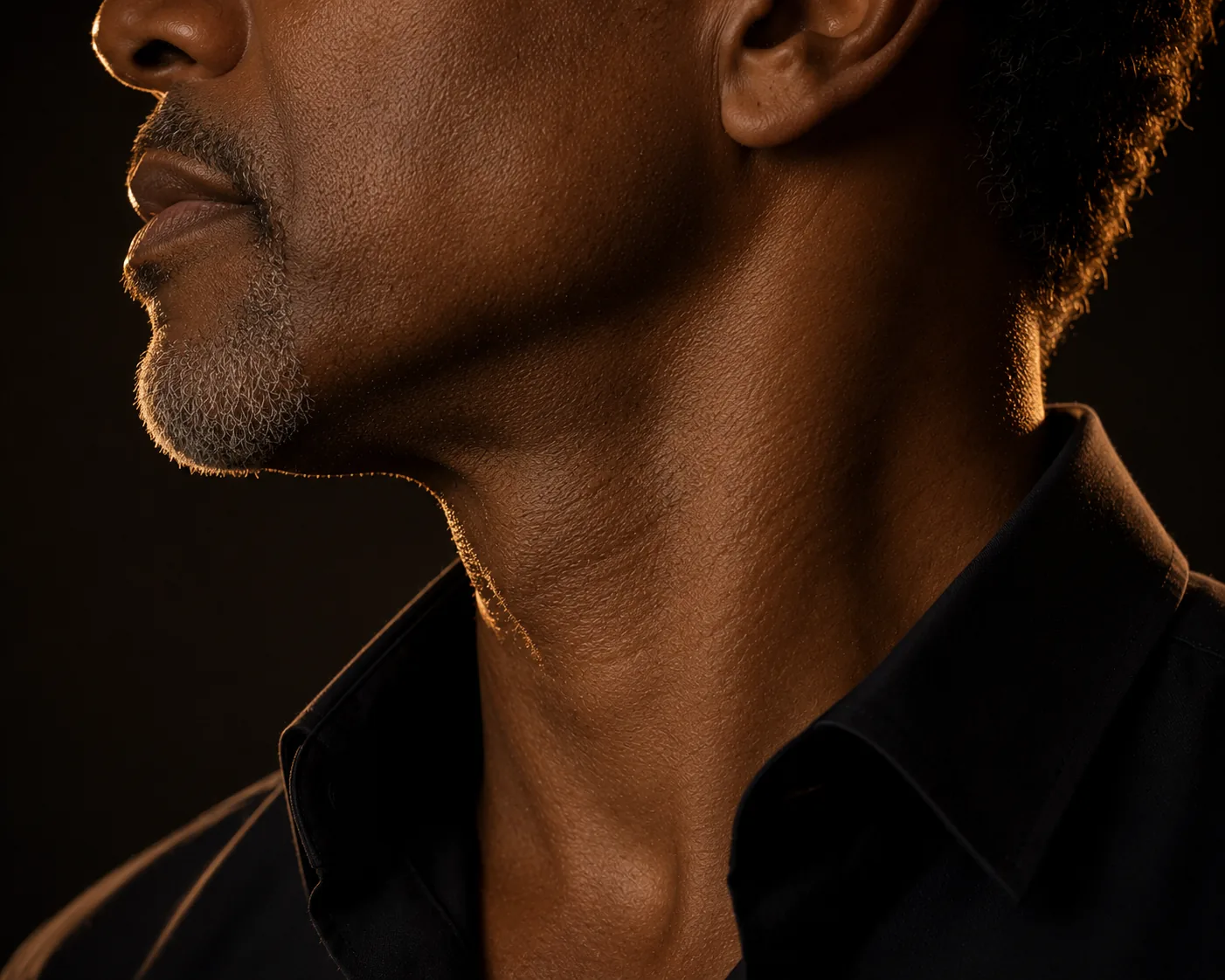Profile of a man's neck and jawline rim-lit in champagne gold against a dark background
