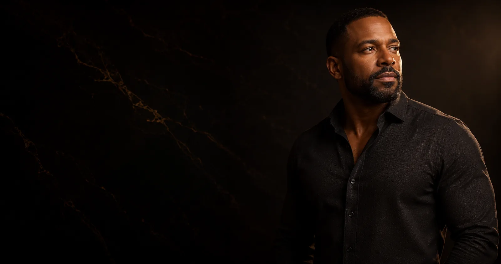 A man in a black shirt standing against dark marble, lit from one side in warm champagne light